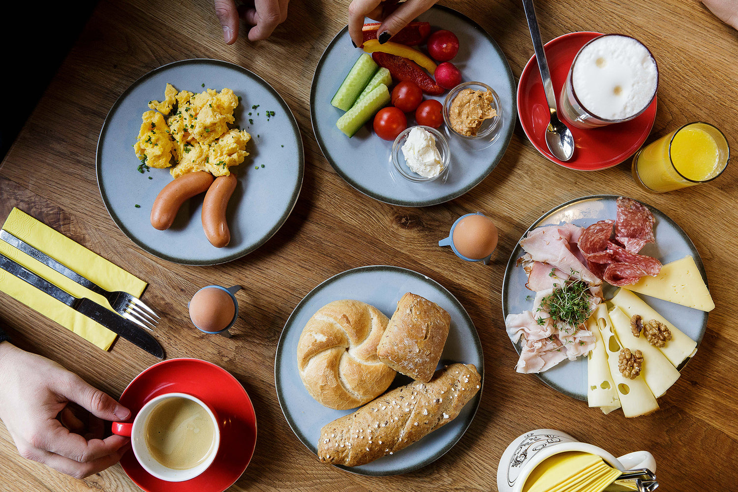 Ein Holztisch auf dem Teller mit Gebäck, Gemüse, Eierspeise, Schinke und Käse sowie zwei Eier, zwei Kaffee und ein Glas Saft stehen.